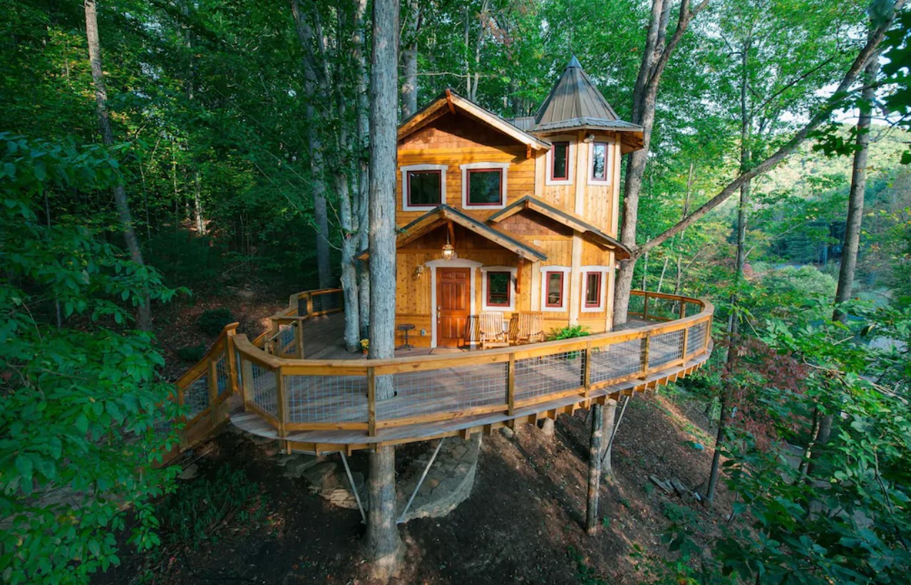 A large treehouse in North Carolina that looks like a small castle. It has a large wrap around deck and is surrounded by trees.