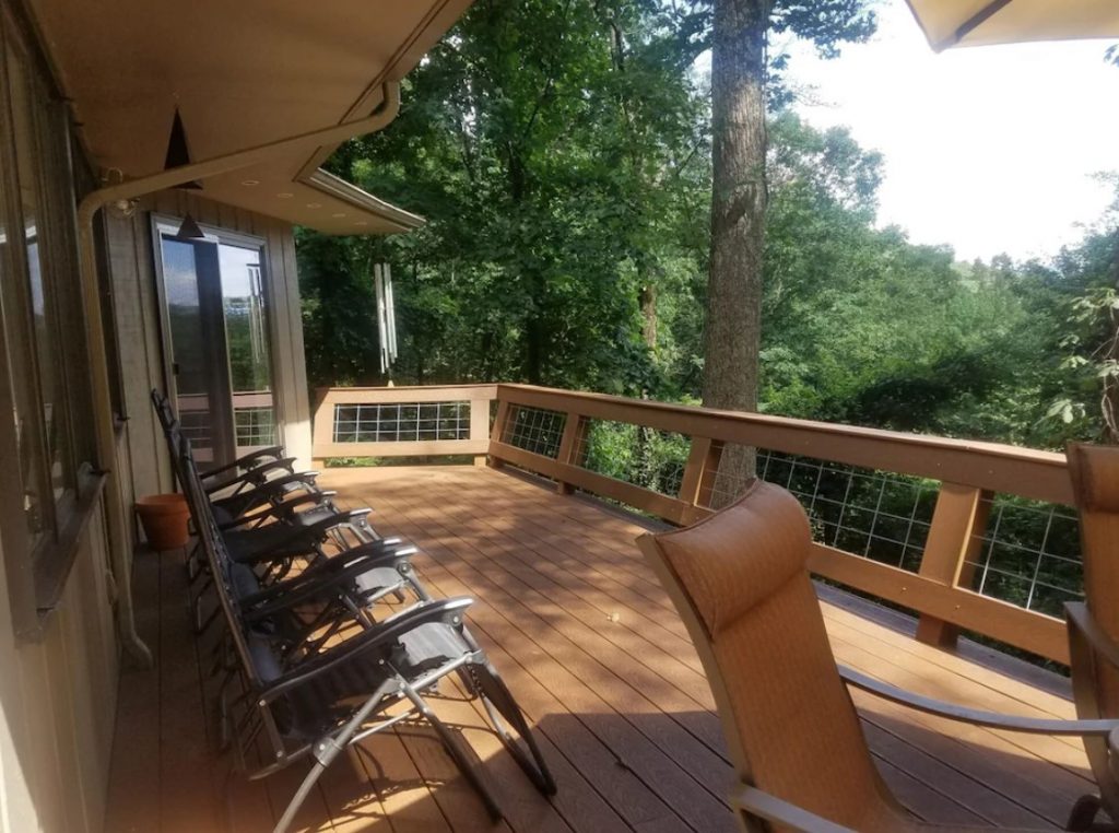 A wooden deck on the side of a cabin that looks out onto the woods. You can see several folding chairs on the deck. 
