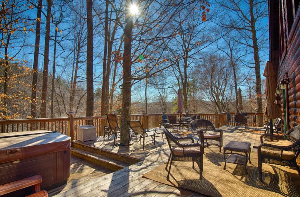 The massive deck of a log cabin treehouse. It has a private hot tub, patio furniture, and a grill. There are trees growing through the deck and views of the woods. 