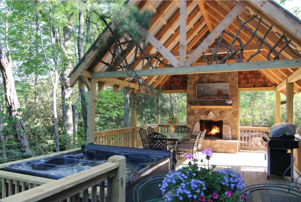 The large deck of one of the best treehouses in North Carolina. It has a covered section, a stone fireplace, a grill, patio seating, and a private hot tub. 