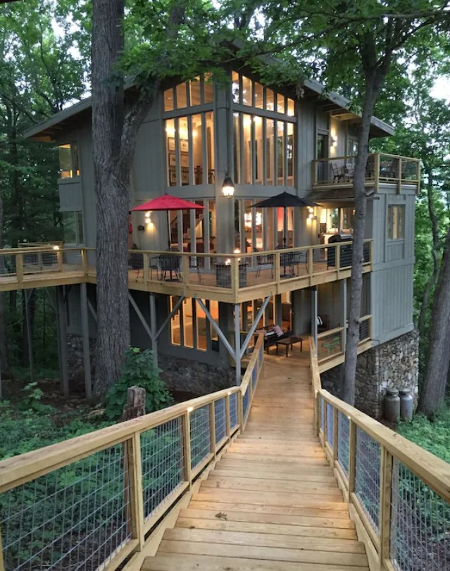 A modern style grey treehouse with three levels of floor to ceiling windows, multiple decks, and a boarded path in the middle of the woods. 