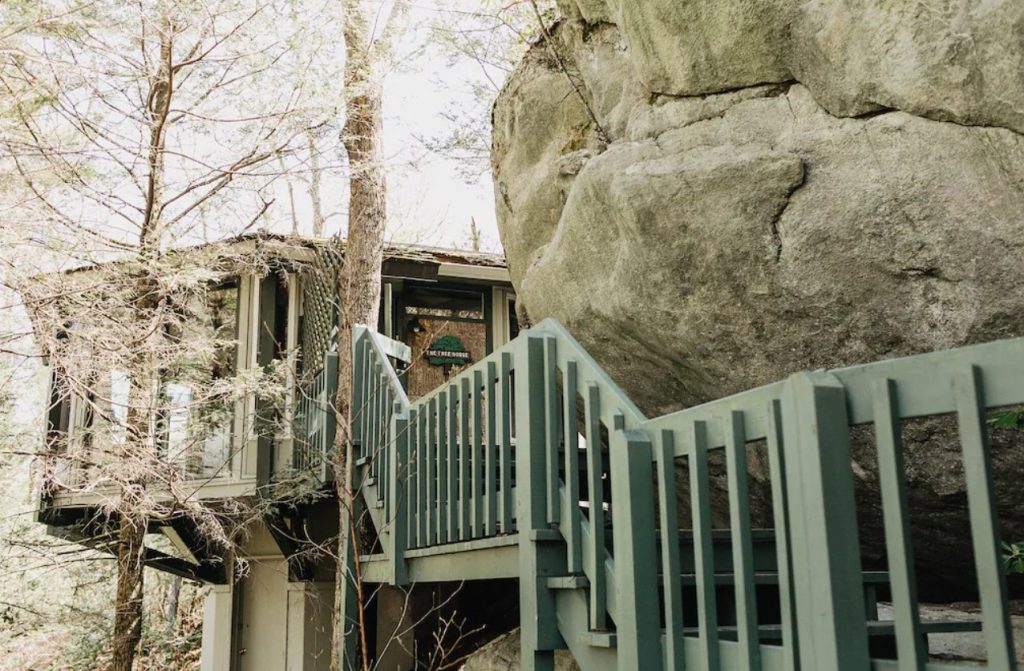 The front entrance of a treehouse on the side of a mountain. You can see a large rock formation right in front of it along a set of stairs. It's one of the best treehouses in North Carolina. 