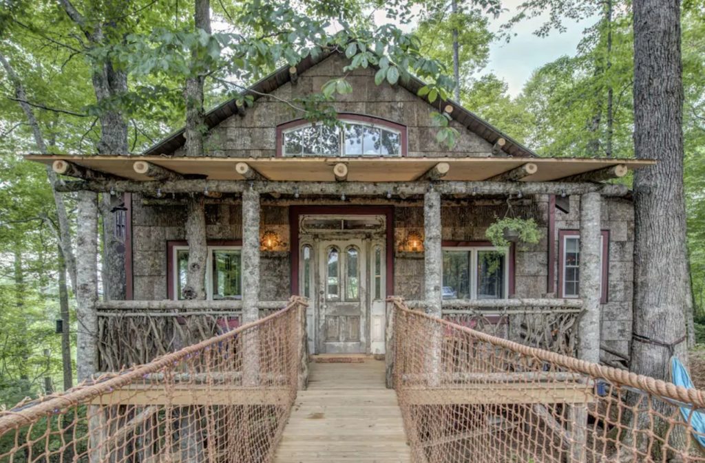 The front of a log cabin treehouse in the woods. It has a large front porch and a bridge with rope leading to the front door. One of the best treehouses in North Carolina. 