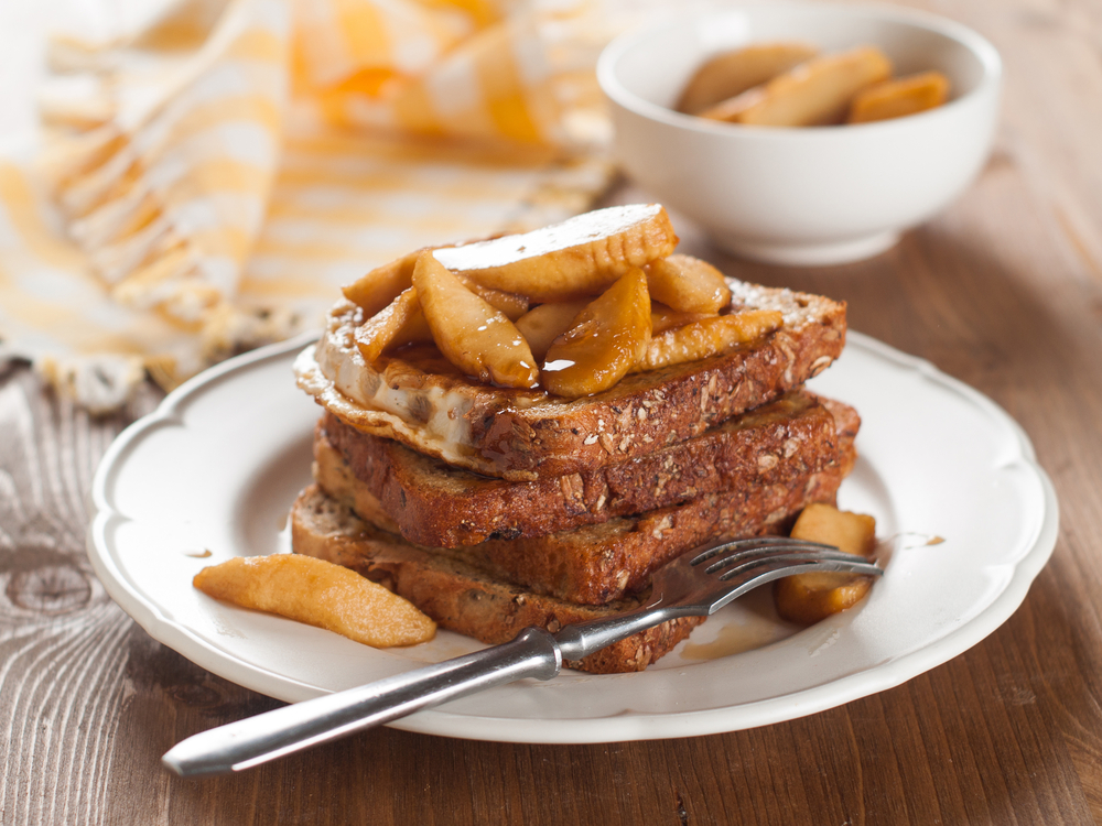 Apple slices sit atop a stack of French Toast, with a fork resting on the plate, similar to the apple French Toast served at Log Cabin Pancake House in Gatlinburg.