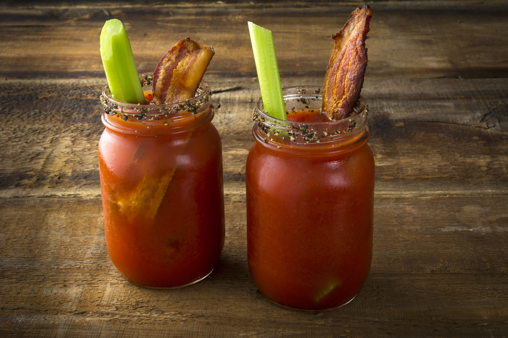 Two Bloody Mary drinks in mason jars, topped with celery and bacon, sit on a wooden table.