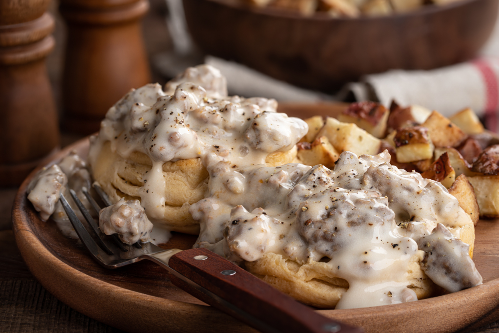 Biscuits and sausage gravy sit on a wooden plate, with potatoes and a fork resting on the plate.