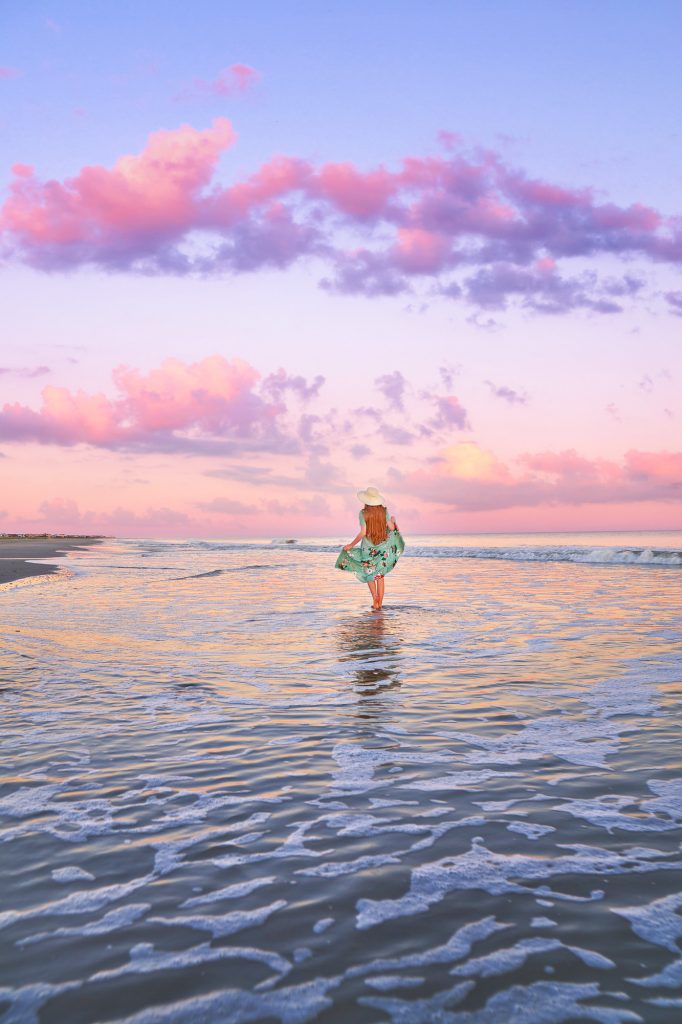 Woman in blue floral dress walking in the water in front of a pink sky.