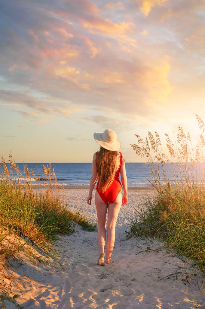 Woman in red bathing suit and hat walking toward the ocean.