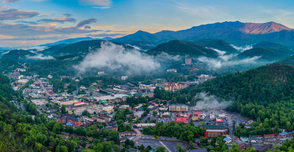An aerial view of the town of Gatlinburg Tennessee. You can see the mountains around it, clouds drifting over the city, and a blue sky. 