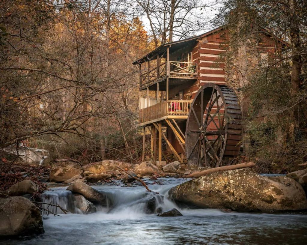 The side view of a two story log cabin with an attached grist mill. It is on the edge of a stream surrounded by woods. Its one of the best cabins in Gatlinburg. 