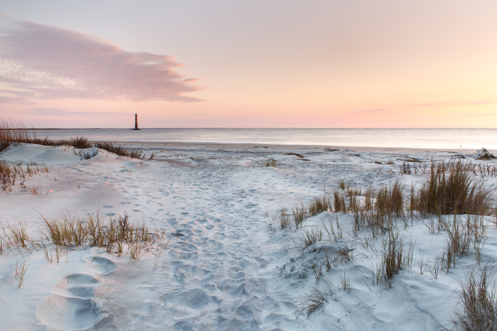 The beautiful white sand of Folly Beach with some beach grass and a lighhouse in the distance. One of the beaches in Savannah. 