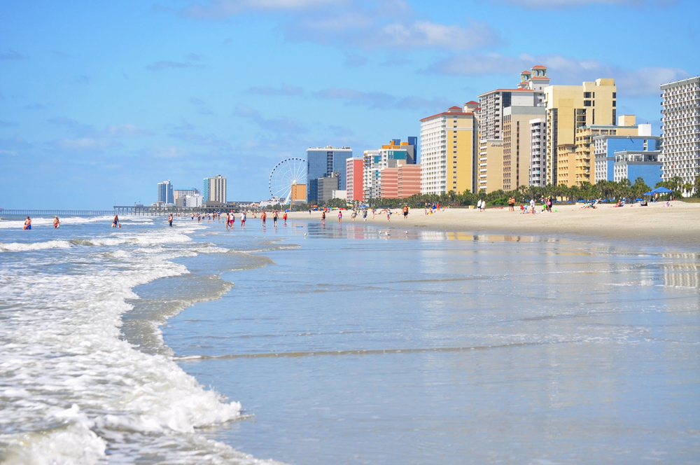 People enjoying the water on a beach with very colourful buildings and a sky wheel.