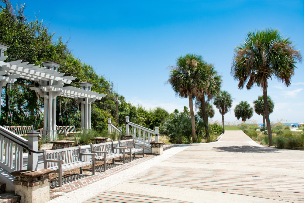 A boardwalk with lush green palm trees, clear skies and white cabanas. 