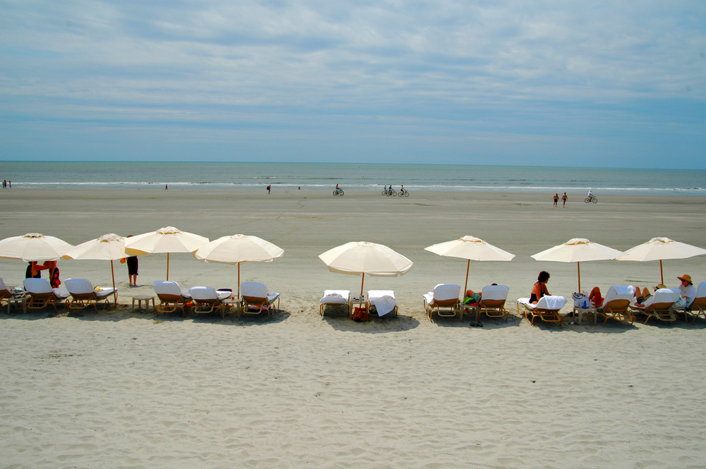 A line of white umbrellas and lounge chairs along a beautiful beach in South Carolina as people bike and walk in the sand.