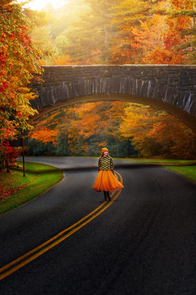 A woman in a sweater and yellow skirt standing in the middle of a road near a stone bridge. You can see the trees changing to orange, yellow, and red. It's one of the best day trips from Asheville. 
