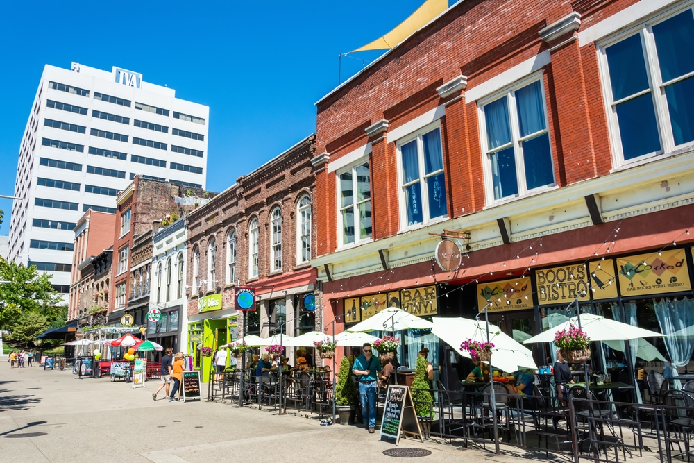 A row of brick buildings that are restaurants. Out front there are patio tables with umbrellas on a sunny day. Its one of the best things to do in Knoxville. 