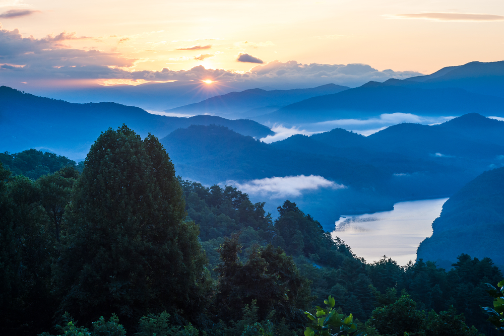 A view of the Great Smoky Mountains as the sun is setting. You can see a lake, clouds in the mountain tops, and trees. It's one of the best day trips from Asheville. 