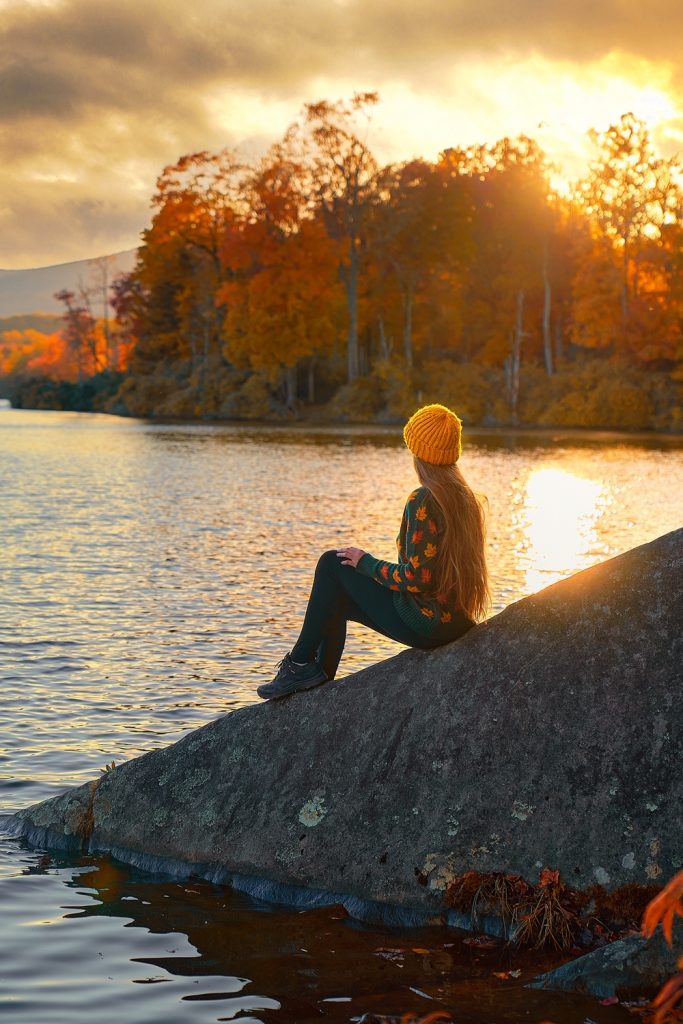 A woman sitting on a large rock by a lake wearing a sweater and knit hat. The trees around the lake are changing to yellow, orange, and red. It's one of the best day trips from Asheville. 