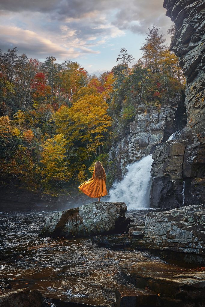 A woman in a yellow dress standing on a rock. She is looking out towards a large waterfall. Around the waterfall there are trees with orange, yellow, and red leaves. 