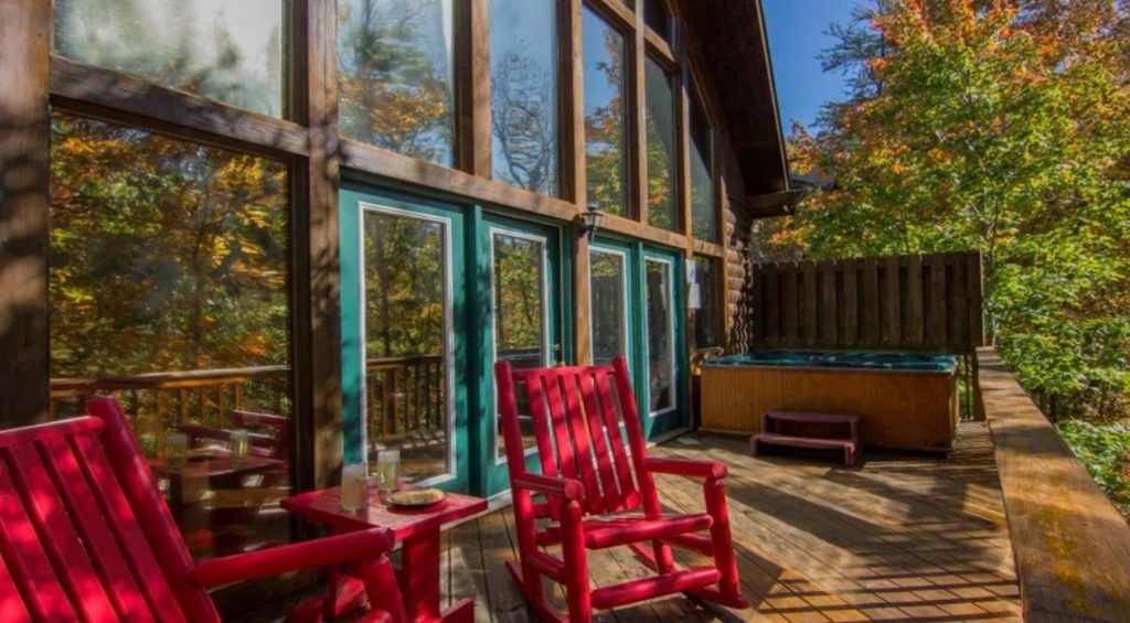 The back deck of a log cabin with large windows and doors. The doors are painted green. There are red rocking chairs and a hot tub on the deck. 