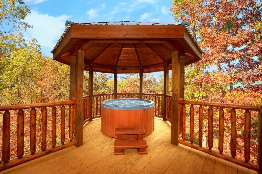 A gazebo on a large deck with a circular hot tub underneath it. Around the deck and gazebo you can see trees with green, yellow, and orange leaves. 
