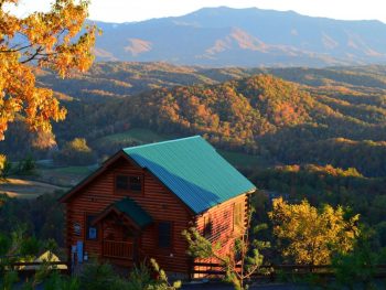 A log cabin with a green roof on the edge of a mountain in the Great Smoky Mountains. Behind the cabin you can see miles of mountains with the trees changing color for fall. It's one of the best cabins in Gatlinburg TN.