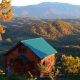 A log cabin with a green roof on the edge of a mountain in the Great Smoky Mountains. Behind the cabin you can see miles of mountains with the trees changing color for fall. It's one of the best cabins in Gatlinburg TN.