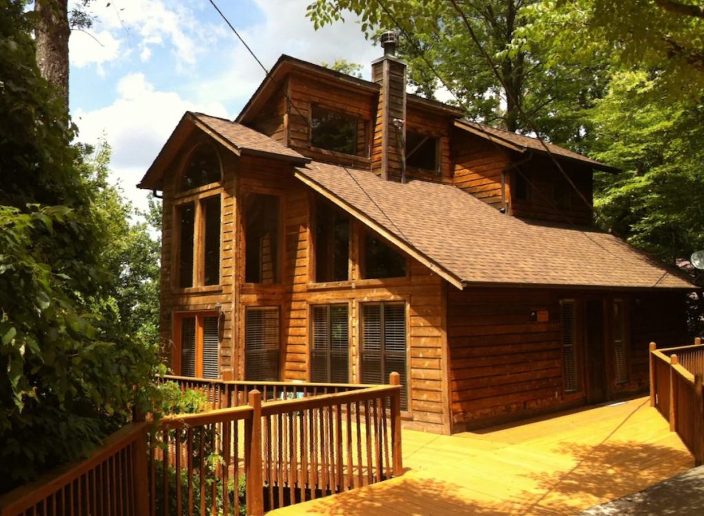A large log cabin with multiple levels and large windows. Leading to the cabin is a large deck and walkway. You can only see trees around the cabin. 