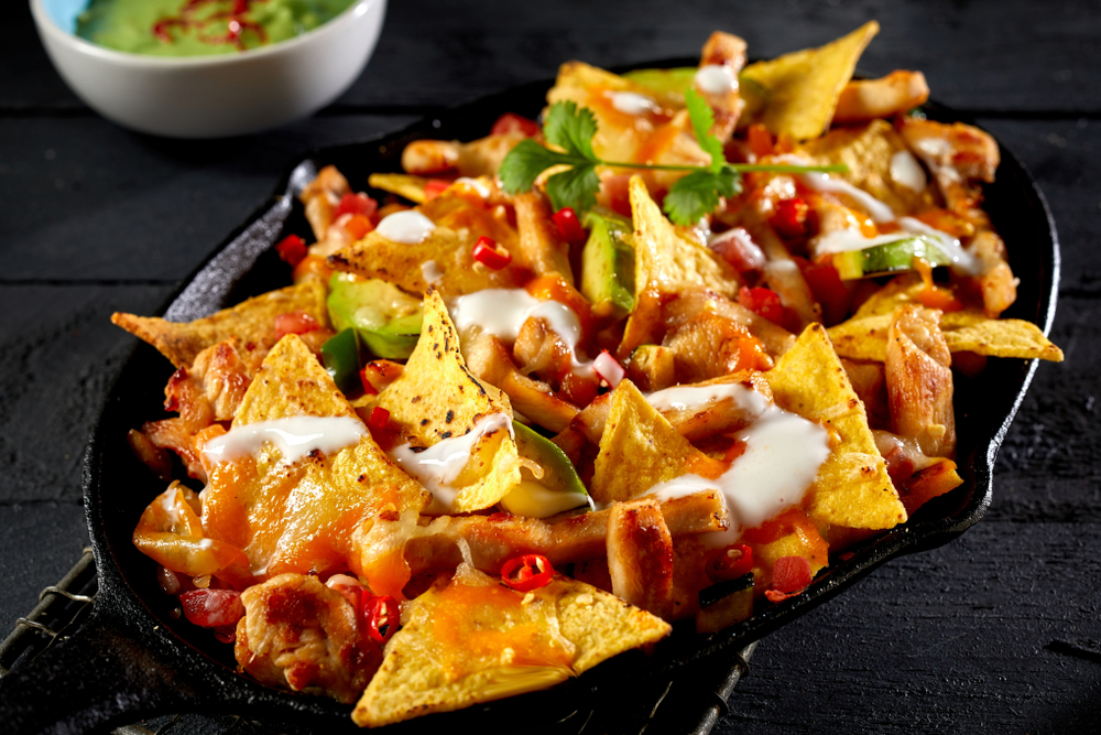 A plate of loaded nachos in a black bowl with a side of guacamole. 