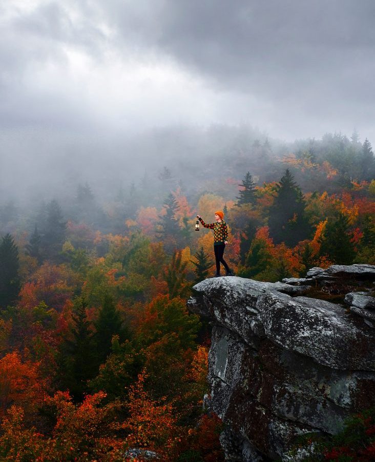 A woman standing on the edge of a rock formation holding a lantern. Behind her there are trees changing to orange, yellow, and red and dense fog. Its one of the best day trips from Asheville.