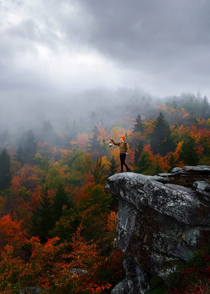 A woman standing on the edge of a rock formation holding a lantern. Behind her there are trees changing to orange, yellow, and red and dense fog. Its one of the best day trips from Asheville. 