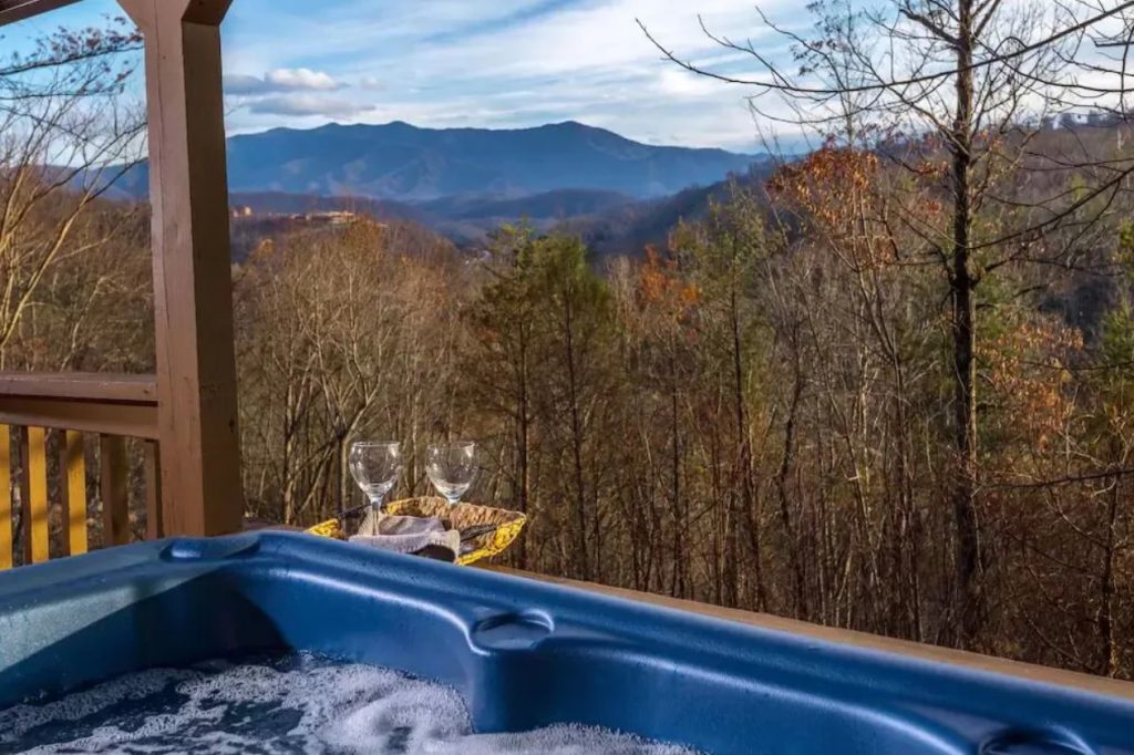 The view of the mountains from the inside of a hot tub on a private deck. You can see the edge of the hot tub, wine glasses, and a wooded area past the deck. 
