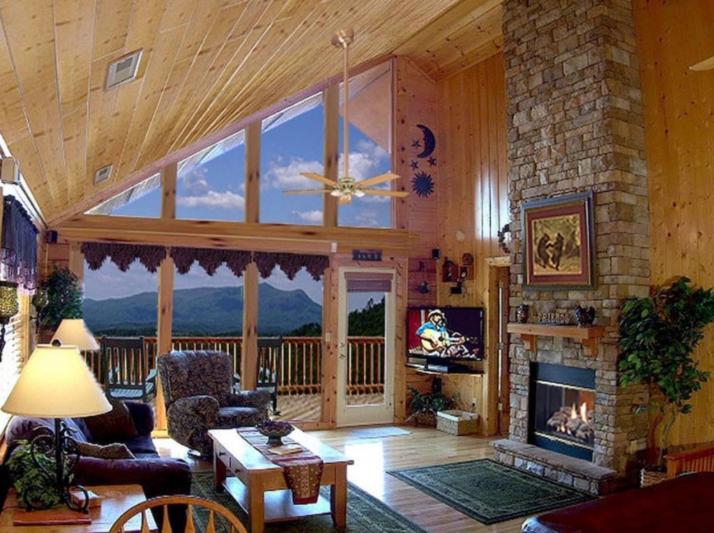 The living room of a cabin with a wall of large windows, a stone fireplace, and living room furniture. From the window you can see the mountains.  