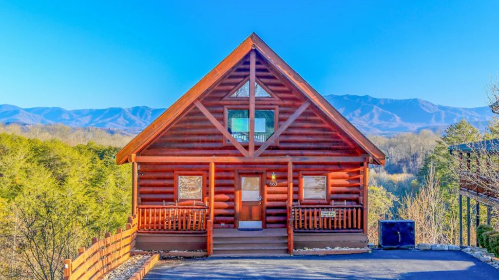 The front of a small classic log cabin. It has a small front porch and a driveway. Behind it you can see unobstructed views of the mountains. 