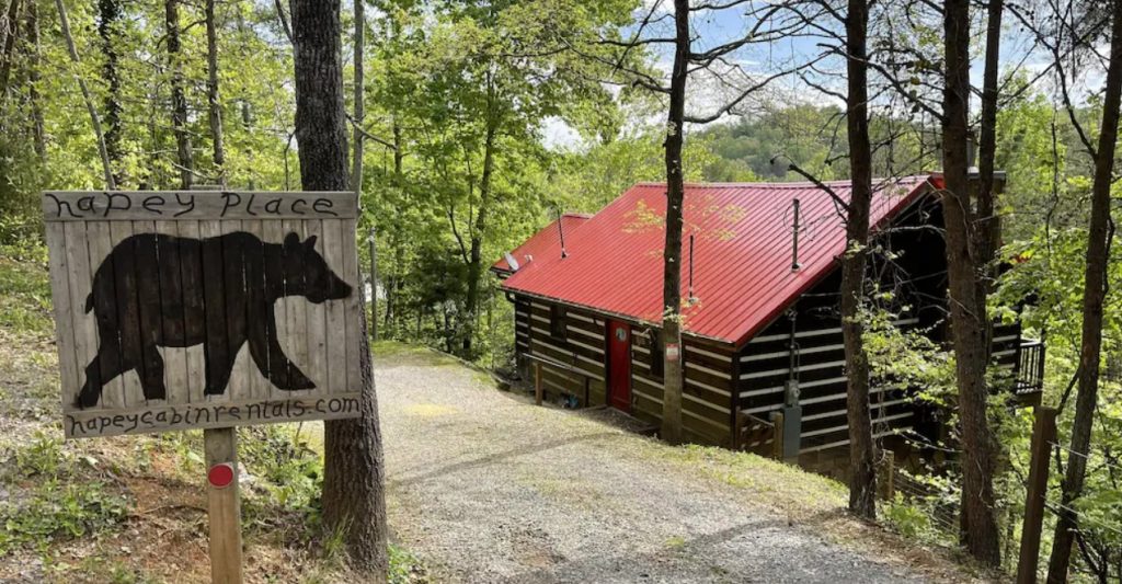 The driveway entrance to a traditional log cabin. There is a sign with a black bear painted on it at the beginning of the drive. Just a little behind it you can see a classic log cabin with a red roof surrounded by trees. It's one of the best cabins in Gatlinburg. 