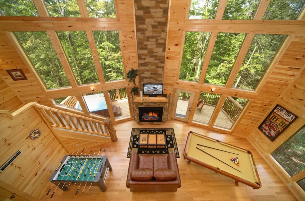 Looking down into a room in a cabin with floor to ceiling windows, a stone fireplace, a couch, a foosball table, and a pool table. From the windows you can see a deck and the woods.