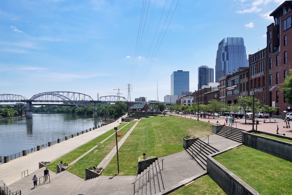 riverfront park with view of a bridge over the water and storefronts to the right