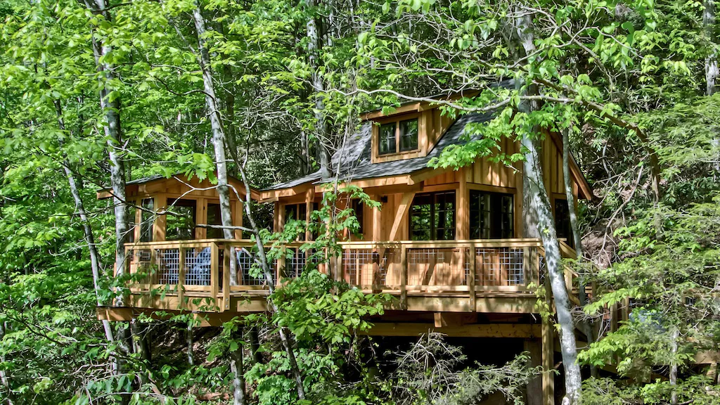 A treehouse surrounded by trees with a large porch and large windows. 