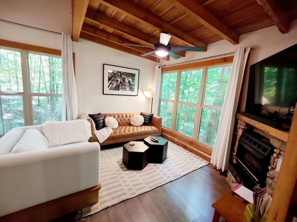 Inside a treehouse with large windows and the trees in the background. The lounge has a stone fireplace and is beautifully decorated in brown and white. 