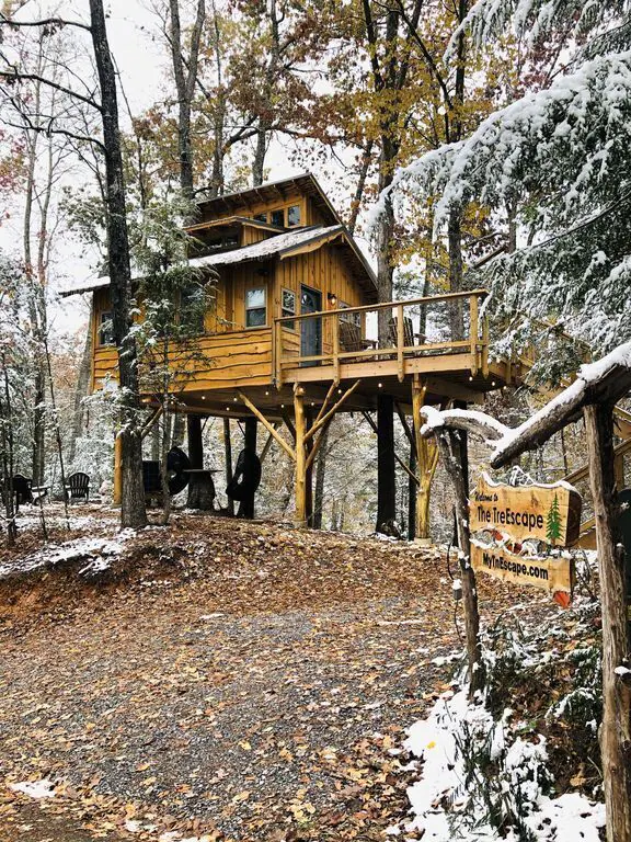 A view from below of a treehouse cabin with chairs underneath and to the side. There is a sign at the front and snow on the ground. 