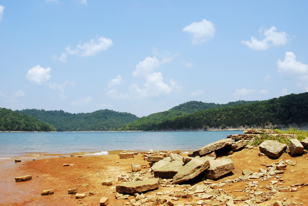 Rocky shores on one of the best beaches in Tennessee, Center Hill Lake, are in the foreground. In the distance are green mountains, enveloping clear blue waters.