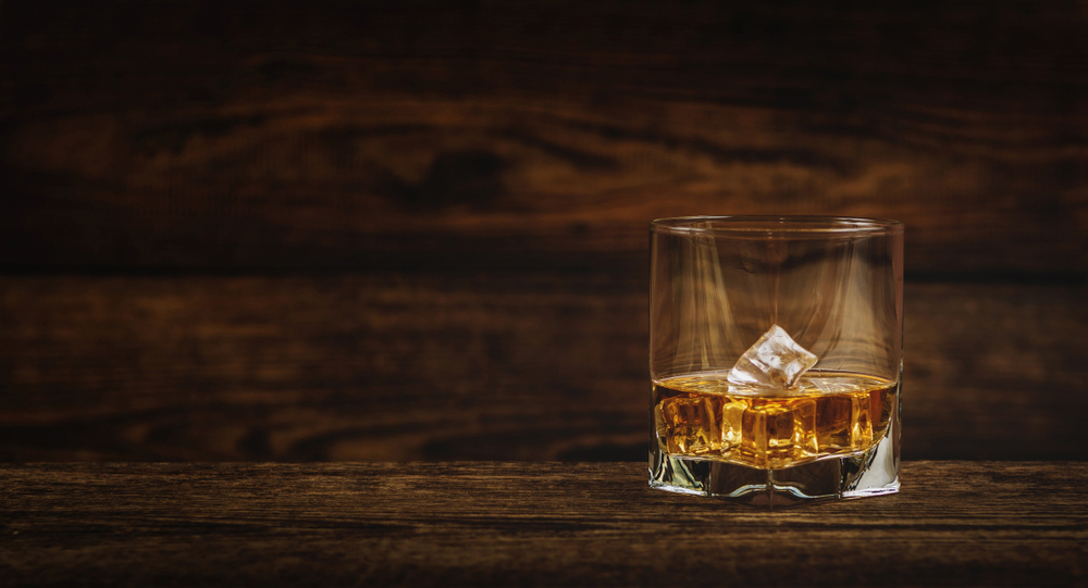 A glass of bourbon with two ice cubes sits on a dark wooden table by itself. The wall behind it is the same dark wood.
