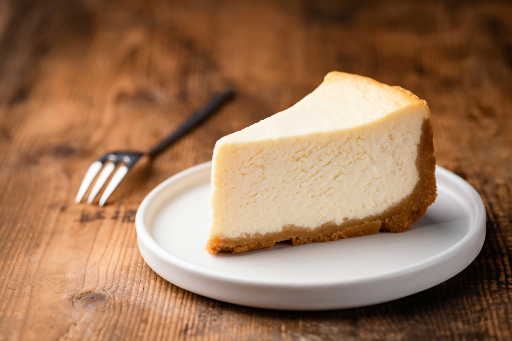 A slice of simple, plain New York style cheesecake is sitting on a plain white plate on a wooden table. A dessert fork sits next to it.