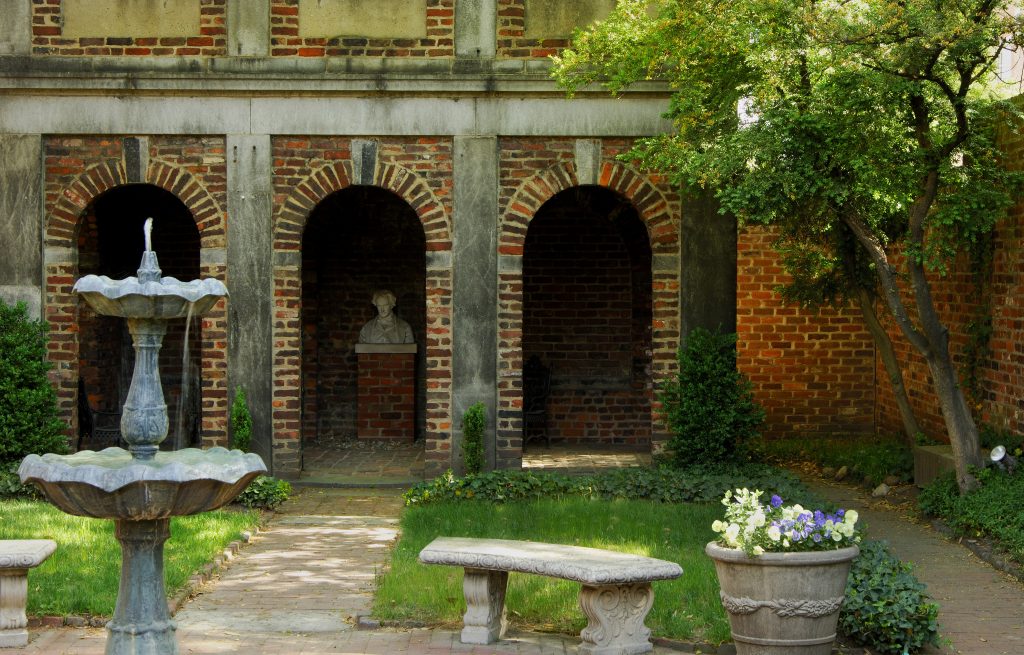 The courtyard at the Edgar Allan Poe Museum in Richmond. You can see a fountain, flowers, grass, trees, and a small alcove with a bust of Poe. 