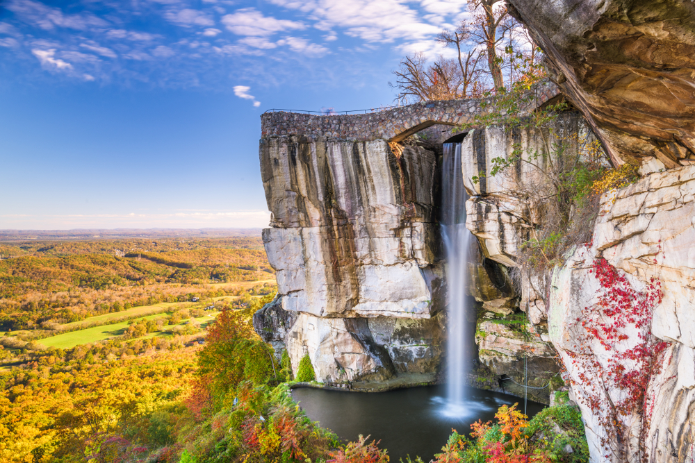 The view of the side of a rocky cliff with a cascading waterfall on the side of it.  Beyond the cliff you can see fields and a forest. Its one of the best hidden gems in Georgia. 