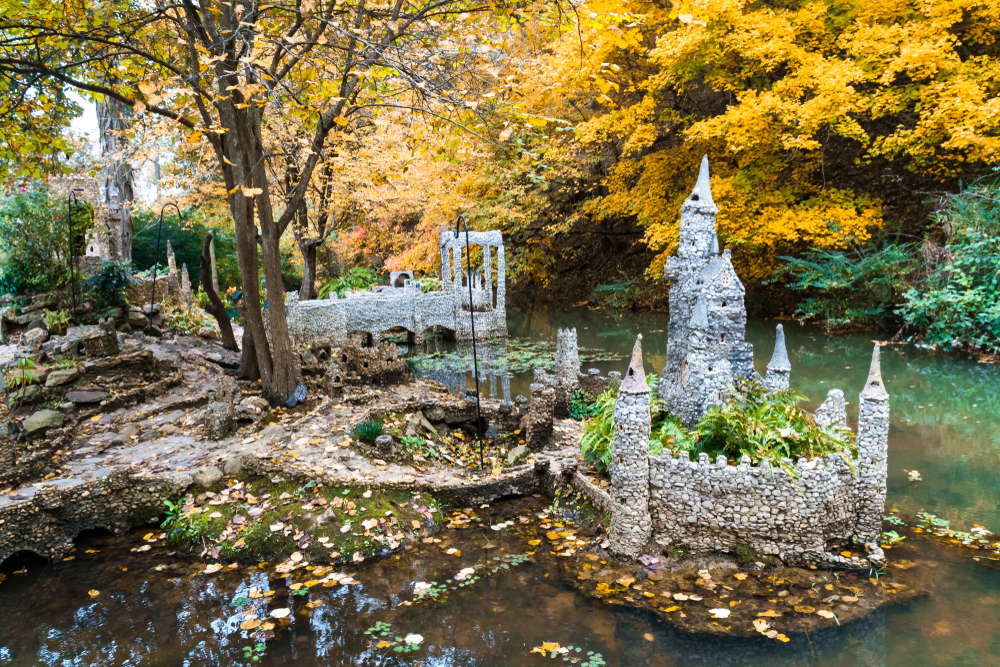 A series of small buildings made entirely of pebbles on the edge of a pond in a garden. There are trees with yellow and green leaves around it. Its one of the best hidden gems in Georgia. 