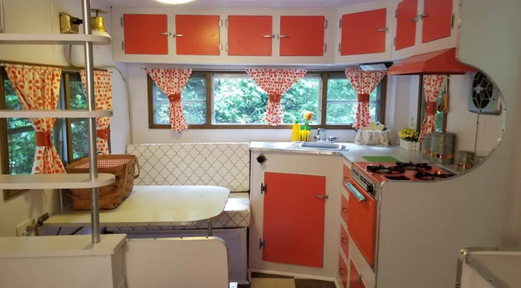 The inside of a vintage camper. There is a small kitchen with an orange stove, orange and white cabinets, and a small dinette. Its a unique place for glamping in North Carolina.