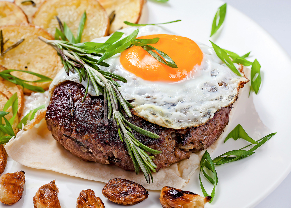 A seared steak with an egg on top. The plate is garnished with roasted garlic fresh rosemary and crispy potatoes.