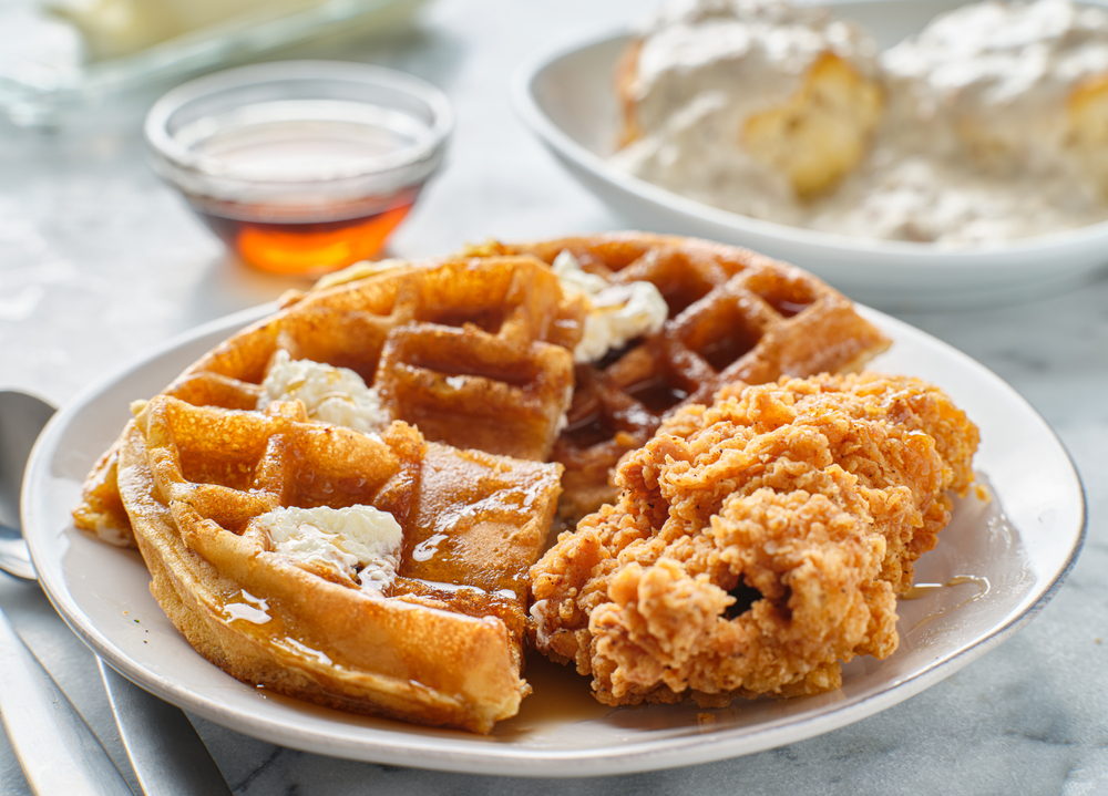 Fried chicken on a plate with slices of waffles, covered in butter and syrup, with a plate of biscuits and gravy in the background.