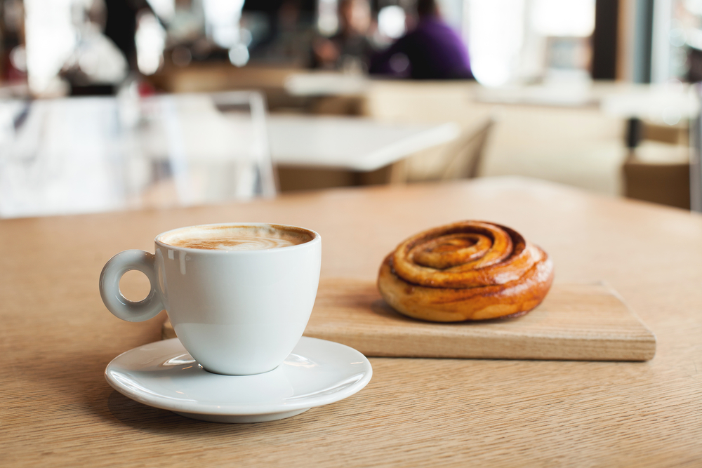 A cinnamon roll sits next to a white cup of coffee on a wooden background, with other tables in the distance.